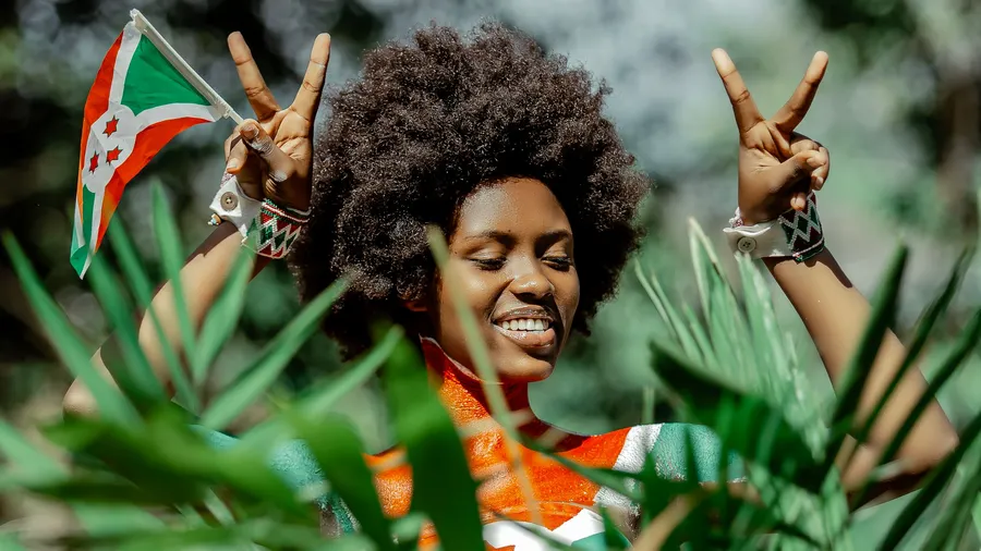Mujer sonriente con peinado afro sostiene la bandera de Burundi entre vegetación y hace señales de victoria