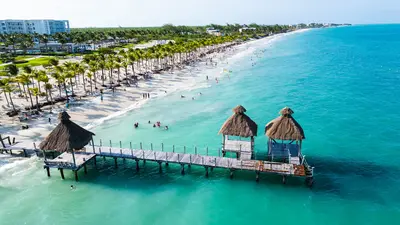 Vista aérea de una playa de arena blanca en Costa Mujeres, con aguas turquesa, palmeras y un muelle de techo de palma