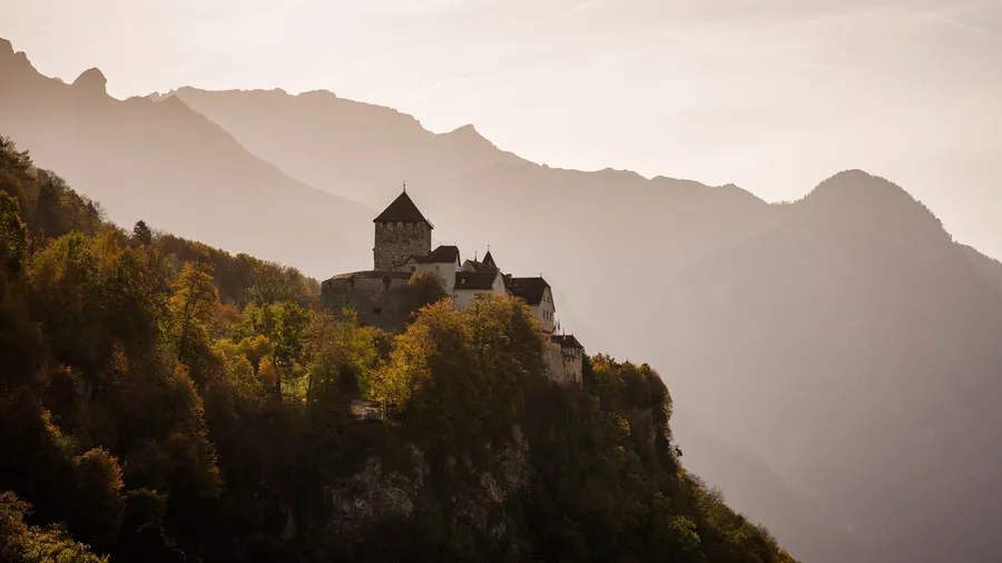 Castillo de Vaduz sobre laderas boscosas, con montañas otoñales al fondo