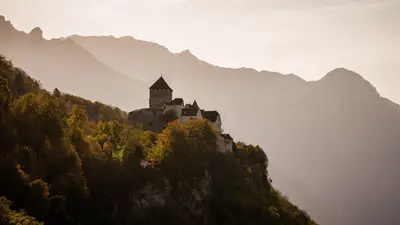 Castillo de Vaduz sobre laderas boscosas, con montañas otoñales al fondo
