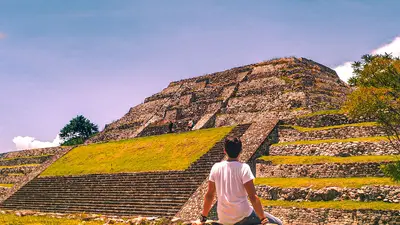 Hombre sentado en un muro de piedra contempla una pirámide histórica en Xochicalco, Morelos