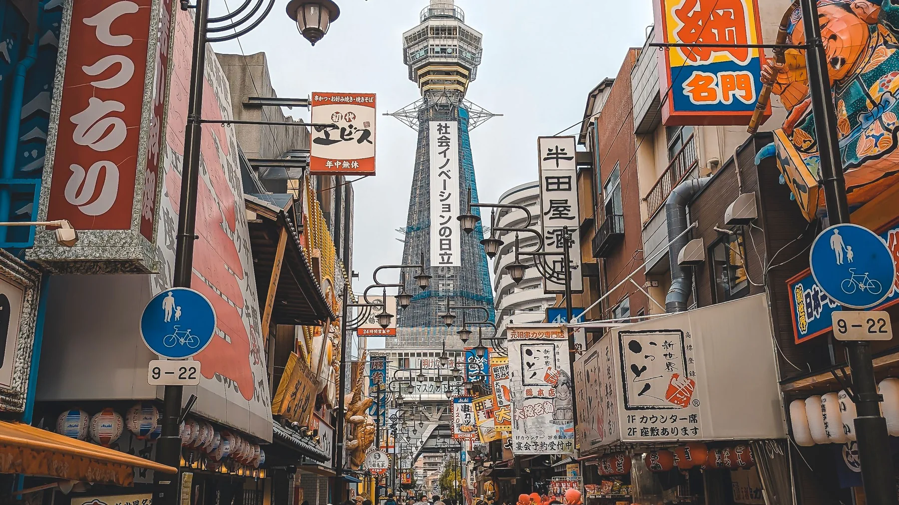 Vibrant urban market street with bustling activity and the iconic Tsutenkaku Tower in Osaka, Japan.