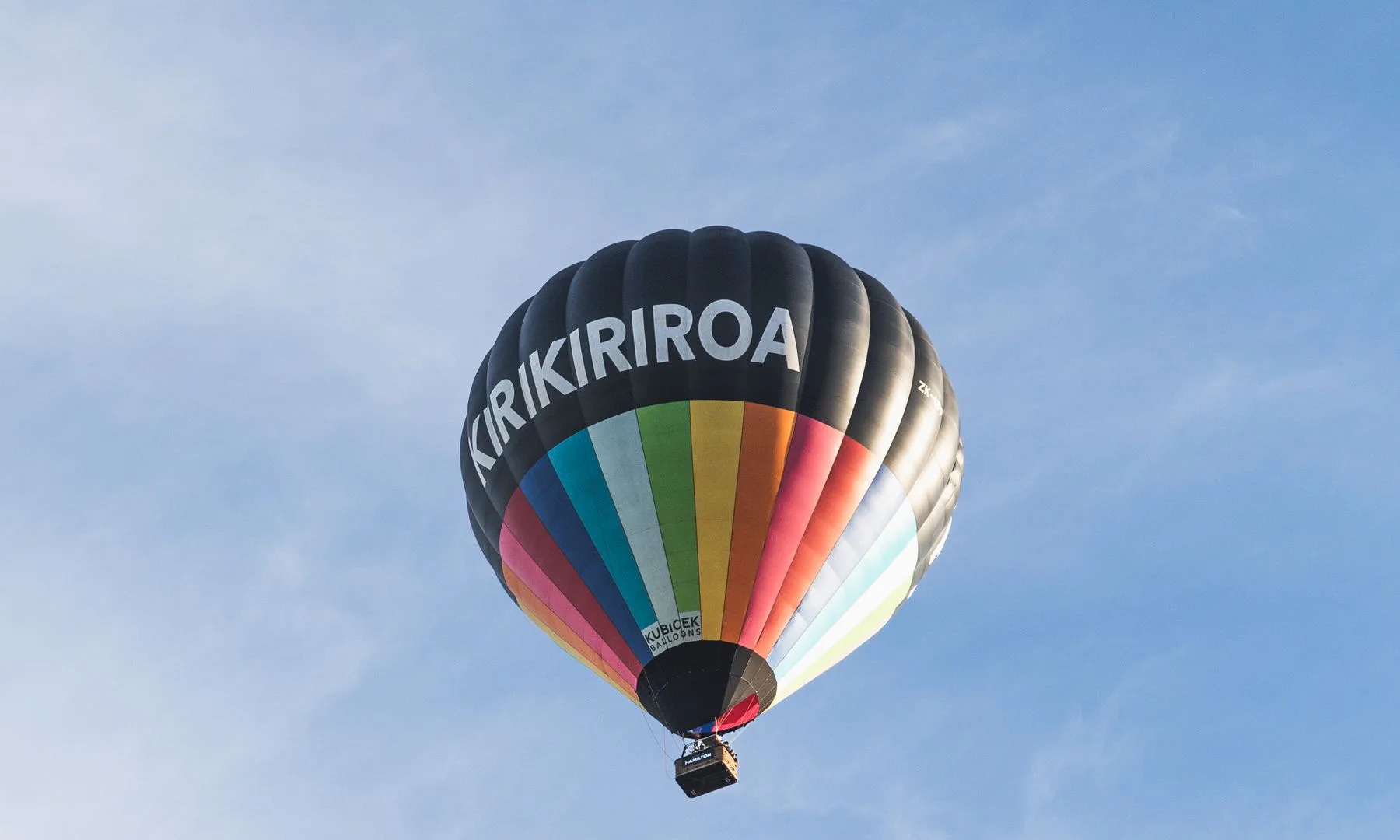 Vibrant hot air balloon with 'KIRIKIRIROA' lettering floats above Hamilton, NZ's clear sky.