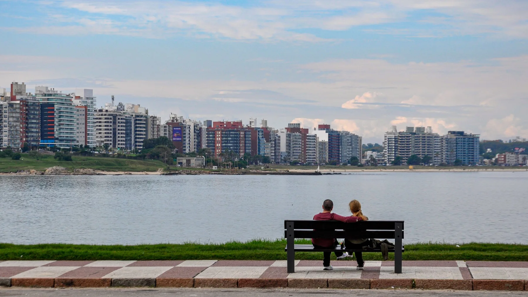 Pareja sentada en un banco frente a la rambla contempla el skyline de Montevideo sobre el Río de la Plata