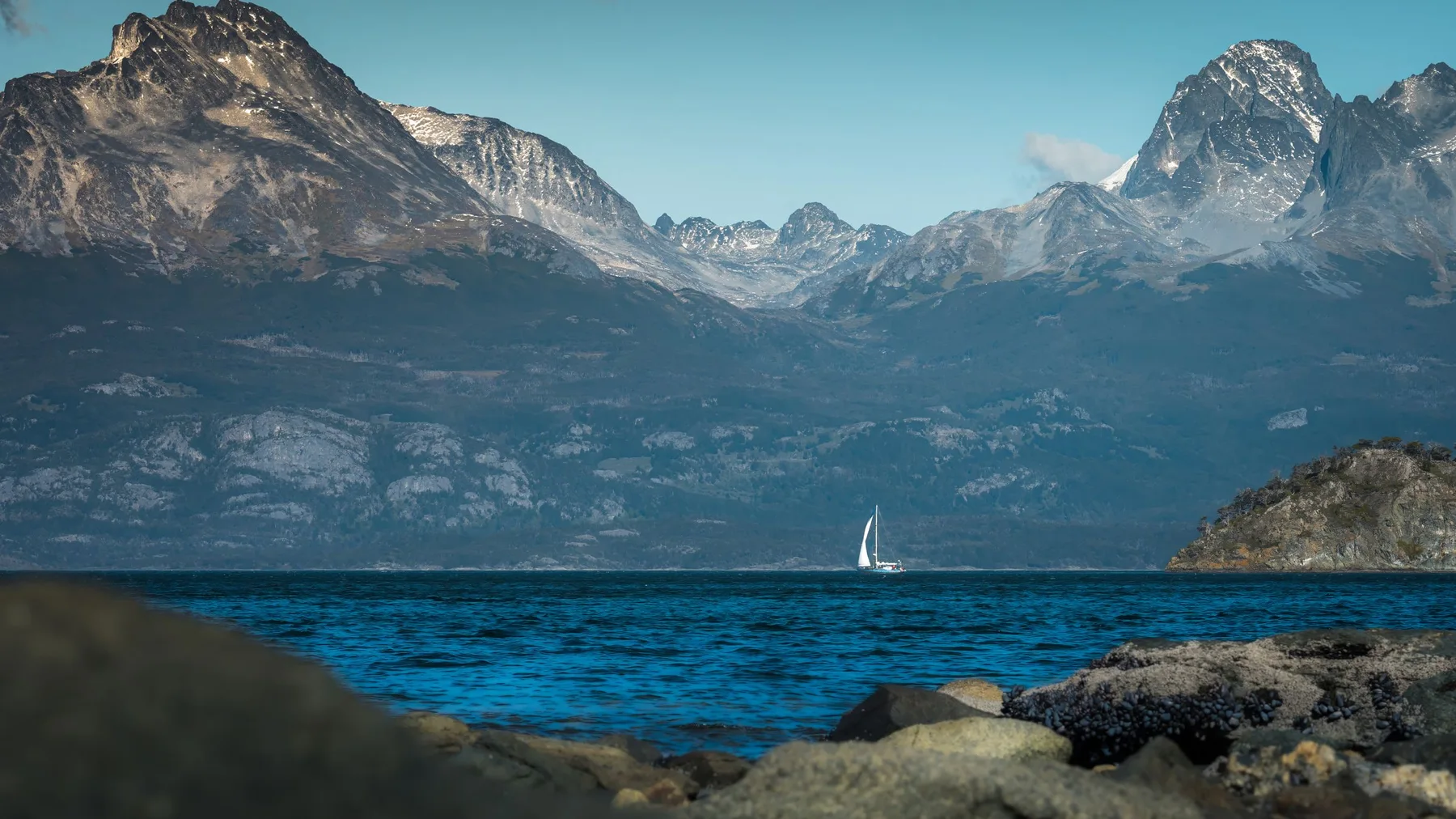 Velero navegando en un lago rodeado de montañas, con Ushuaia y la cordillera de los Andes de fondo