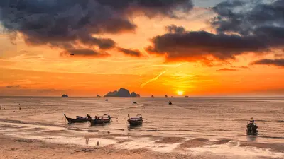 Atardecer dorado sobre una playa de Krabi, con barcos tradicionales y nubes dramáticas